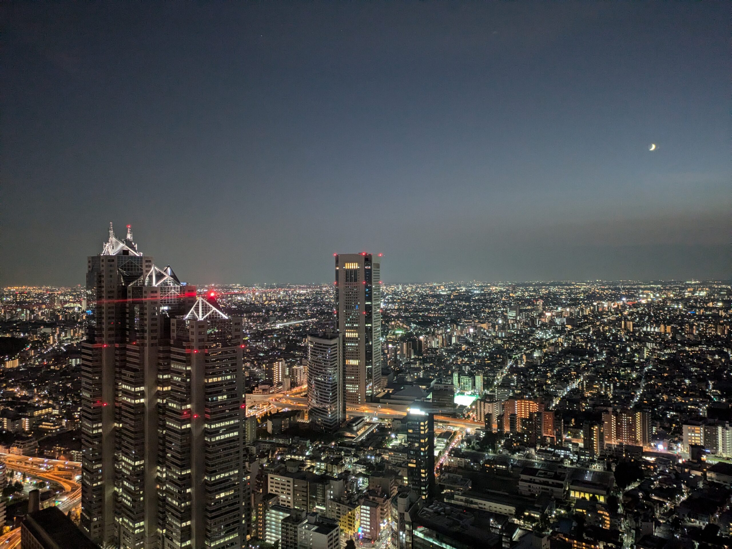 Landscape image of Tokyo at night from an tall observation tower. Crescent moon is visible in the top left corner.