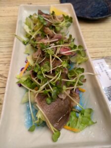 A rectangular plate features thin slices of seared beef garnished with vibrant microgreens and served on a bed of mixed greens. The beef is lightly cooked and pink in the middle, with a fresh and crisp presentation. The dish is accented with a lemon wedge and colorful vegetables.