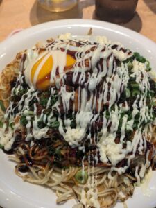 A bowl of noodles topped with a rich, golden egg yolk and a generous drizzle of white mayo and dark savory sauce. The dish is garnished with chopped green onions, which are scattered across the top, giving a fresh and hearty look. The noodles have a stir-fried texture, with visible caramelization in places.