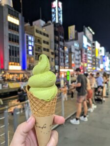 A cone of matcha-flavored soft serve ice cream held up in front of a lively, brightly lit street scene at night. The ice cream is a rich green color, with a perfectly swirled top. The cityscape in the background is illuminated with neon signs and bustling with people.