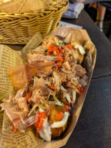 Dough balls with octopus inside and a variety of bright red and green sauces and toppings on top. Served in a compostable tray on an outdoor patio restaurant in Osaka, Japan. 