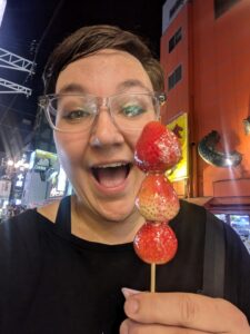 A smiling person (Christine Cuellar, blog post author) holds a skewer with three large, shiny strawberries in front of a brightly lit street at night. The strawberries are glazed, giving them a glossy appearance, and Christine's expression shows excitement. The vibrant city lights and signs create an energetic background scene.