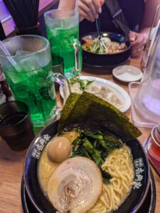 A bowl of Tokyo ramen with a smoked softboiled egg, nori (seaweed), and a round slice of pork on top. A bright green melon soda in a large beer mug is next to the bowl. 