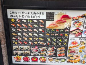 Large backlit poster menu with many pictures and descriptions of sushi. Text is in Japanese, but because the pictures are so specific, the menu is understandable regardless of the viewer's primary language. 