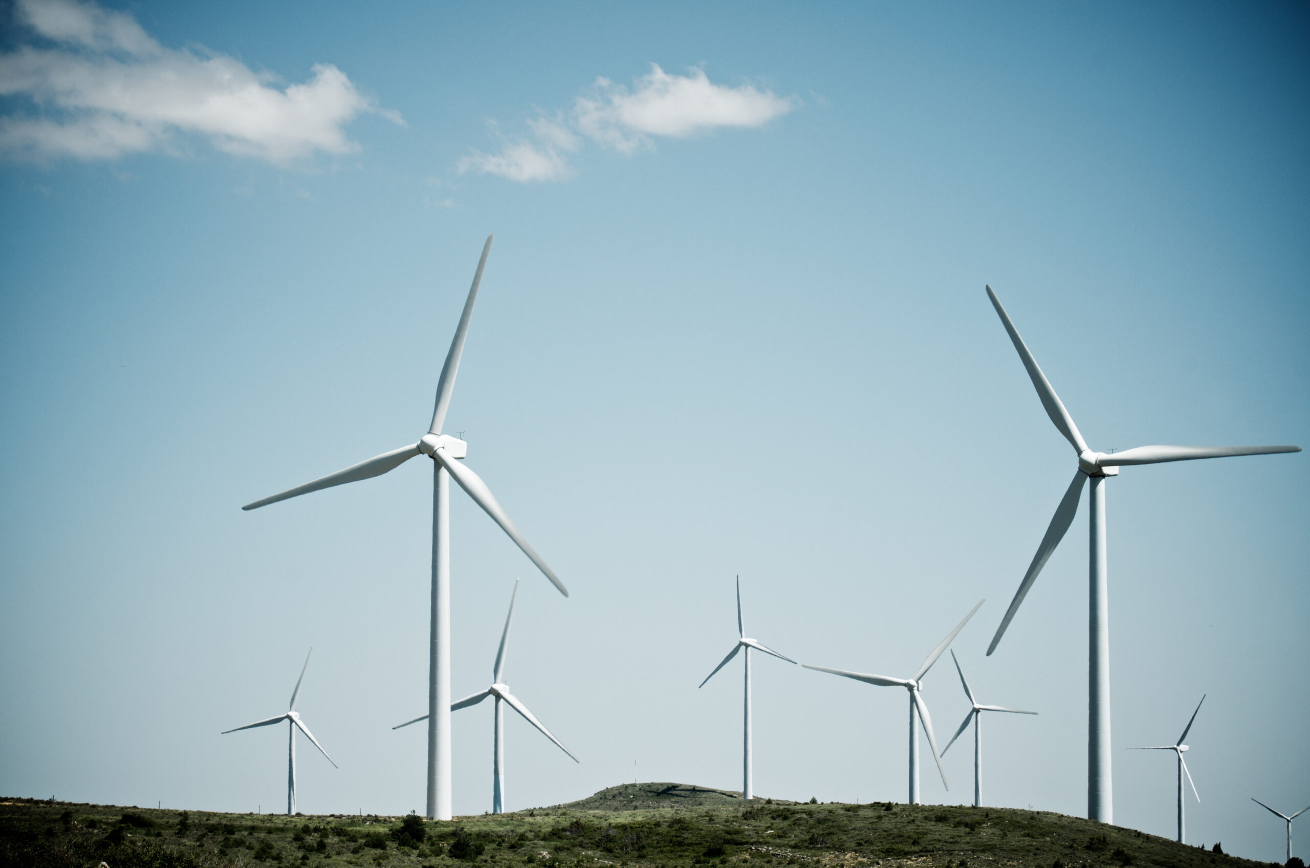 This image shows several large wind turbines standing tall on a grassy hillside under a clear blue sky. The turbines' long blades are positioned to harness the wind's power, symbolizing purposeful, sustainable energy generation. Unlike a hamster wheel spinning in circles aimlessly, these turbines turn with clear intent, converting wind into valuable electricity. The image evokes a sense of efficient, directed movement—spinning with a purpose.