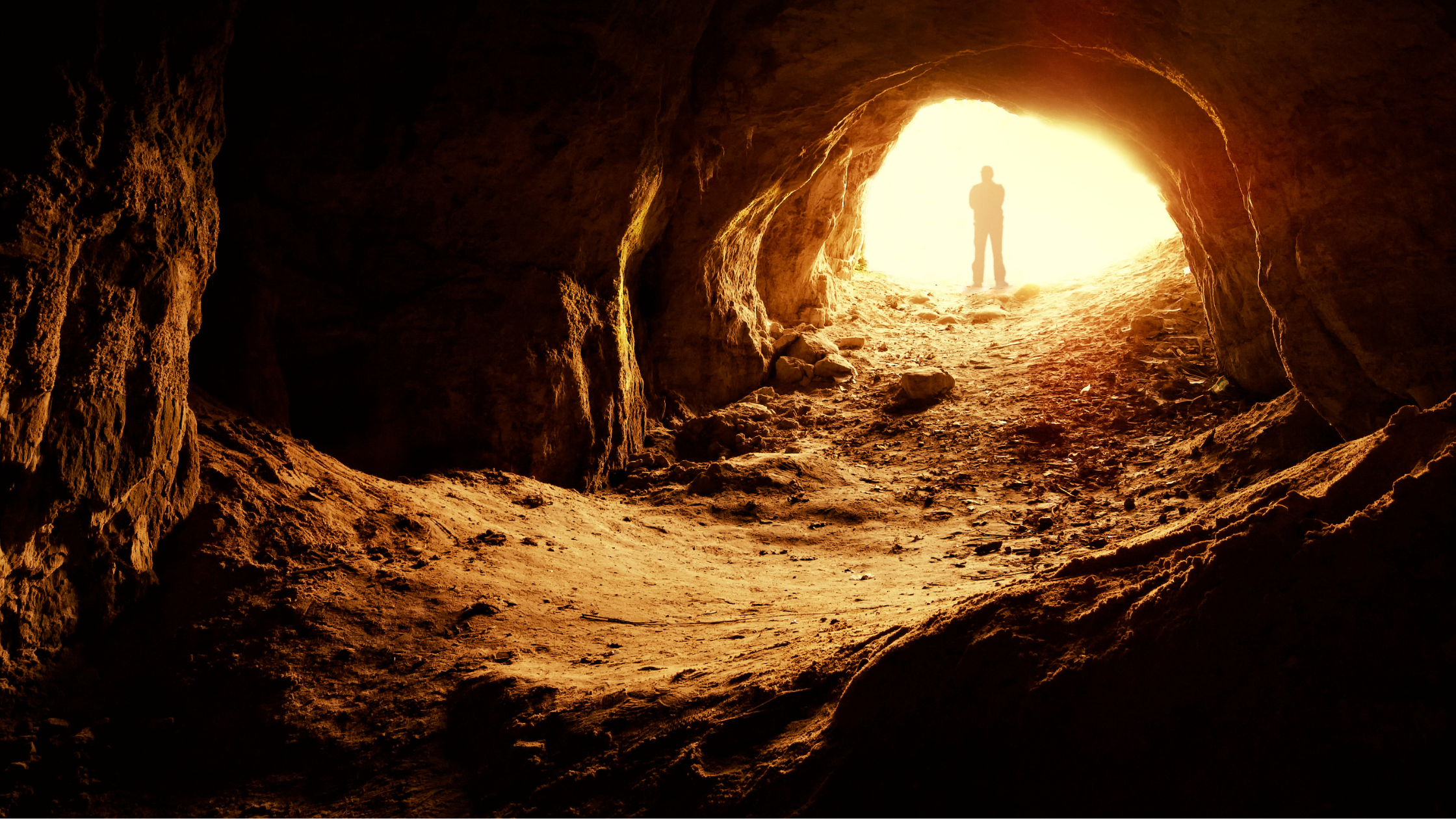 This image shows the inside of a dark cave with rocky walls, leading toward an opening where bright light floods in. At the cave's exit, a silhouetted figure stands in front of the glowing sunlight, casting a mysterious and dramatic atmosphere.