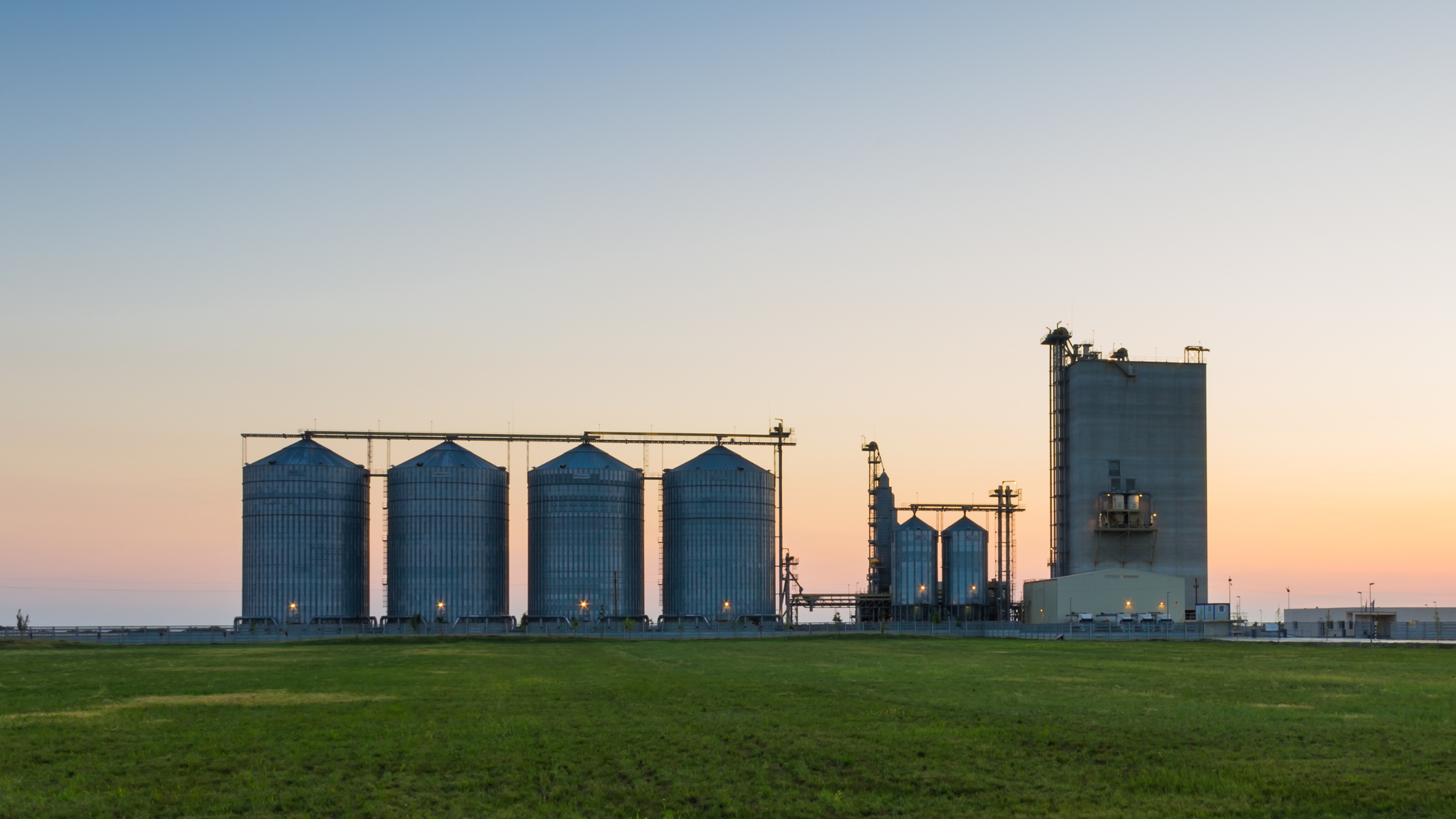 This image shows a wide landscape featuring several large grain silos and an industrial structure against the backdrop of a sunset. The silos are aligned in a row, with the larger industrial building on the right. The sky is softly lit in shades of orange, pink, and blue as the day transitions to dusk. The foreground is a green grassy field, and some lights on the silos and industrial building are turned on, signaling the end of the day. The scene conveys silos and disconnect in content departments within an organization (marketing, technical, support, and knowledge base).