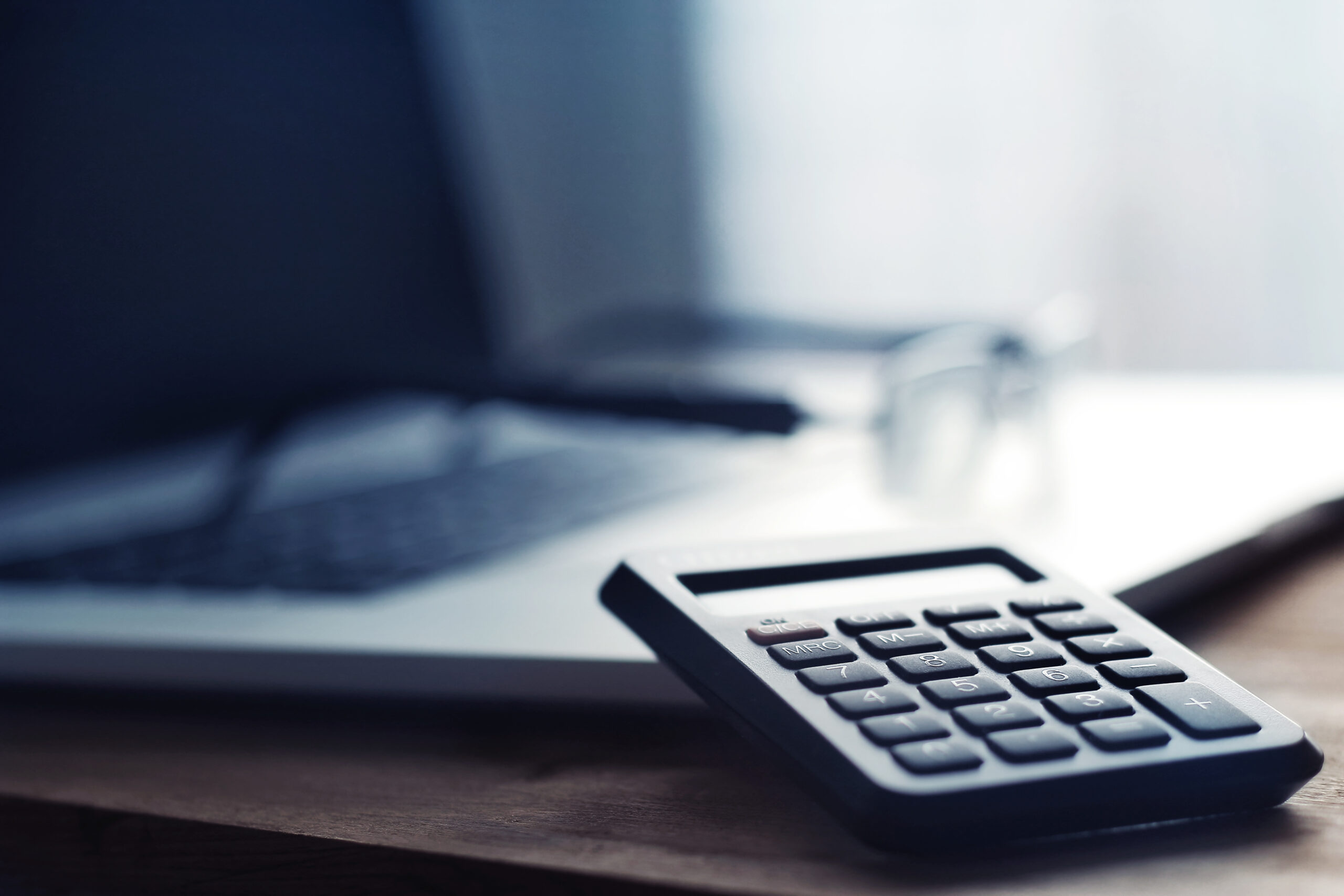 A close-up photograph of a calculator resting on a wooden desk, with a laptop and a pair of glasses in the blurred background. The image has a cool-toned color palette and a shallow depth of field, emphasizing the calculator in the foreground while the background elements appear softly out of focus.